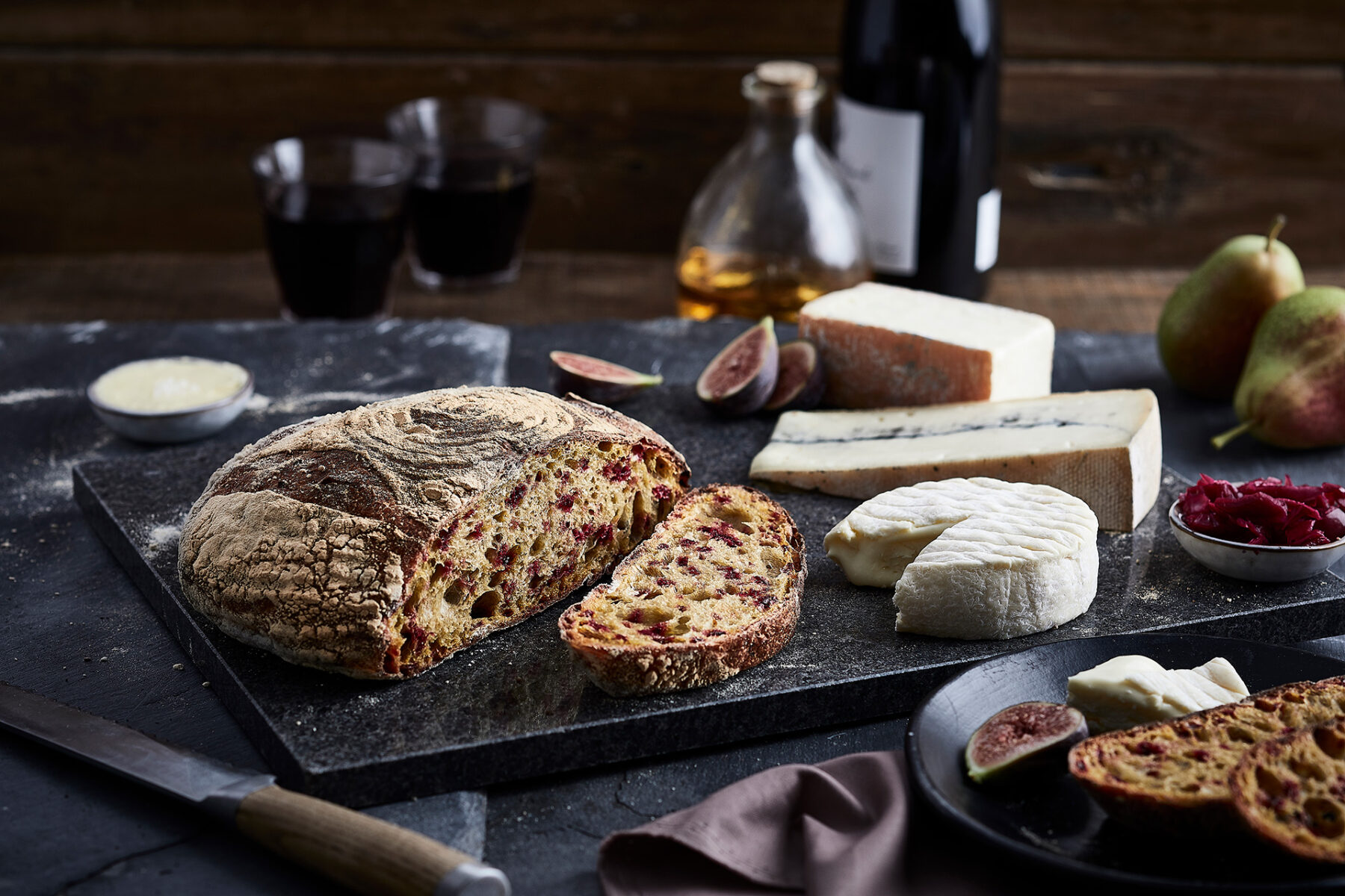 Image of an oval shaped loaf of Beetroot sourdough on a black cheeseboard surrounded by figs and cheeses