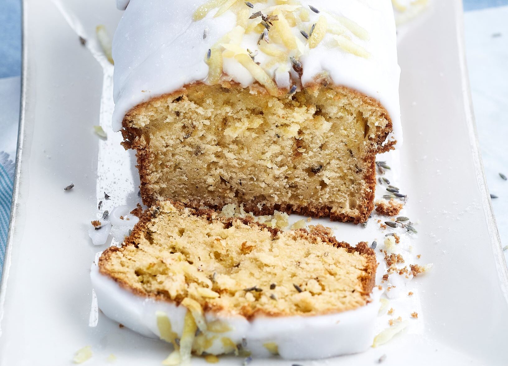 Image of a Lemon and Lavender Cake in a loaf shape and white icing over the top, the first piece has been sliced and is lying in front of the cake.