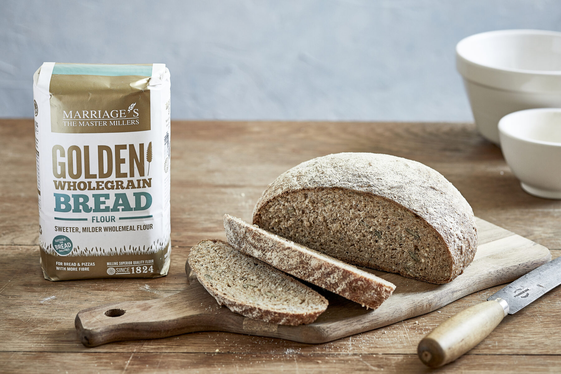 Image of Marriage's golden wholegrain bread flour in a whit and gold packet next to an oval shaped brown loaf of bread